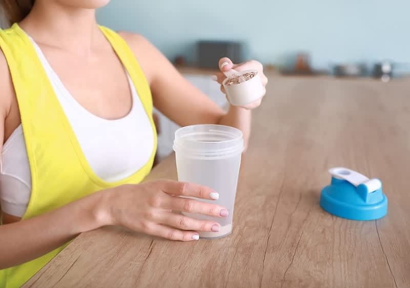 Donna prepara frullato proteico con misurino e shaker su tavolo di legno