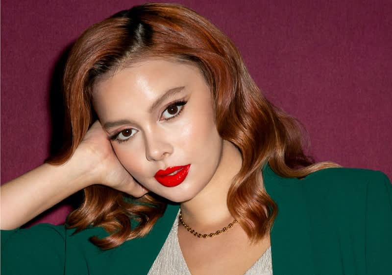 Donna con capelli ondulati castani e blazer verde su sfondo rosso