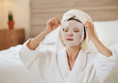 Persona in accappatoio bianco con asciugamano in testa applica maschera viso in tessuto