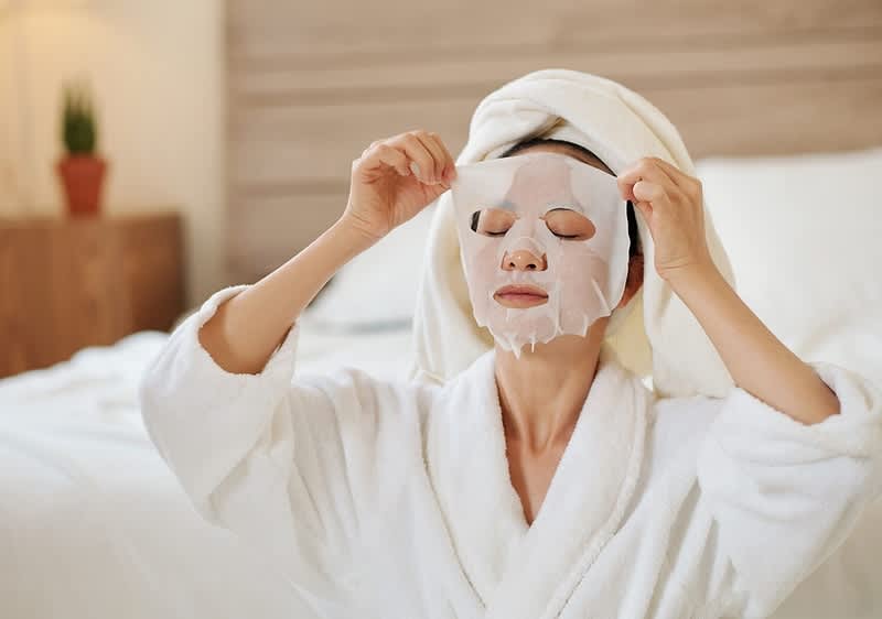 Persona in accappatoio bianco con asciugamano in testa applica maschera viso in tessuto