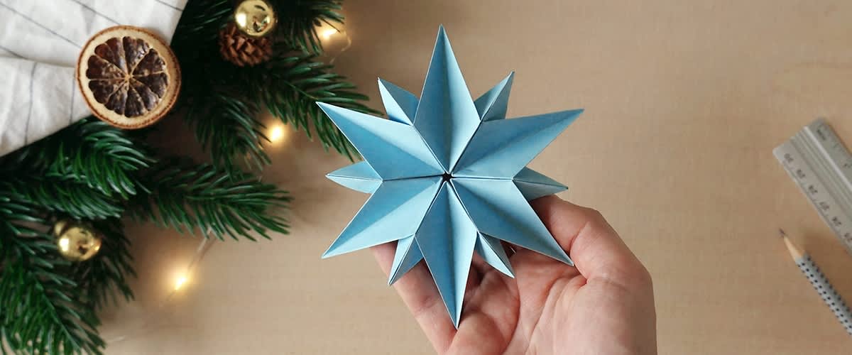 Una mano tiene un origami a forma di stella realizzato con carta azzurra su sfondo neutro con albero di Natale con addobbi e luci