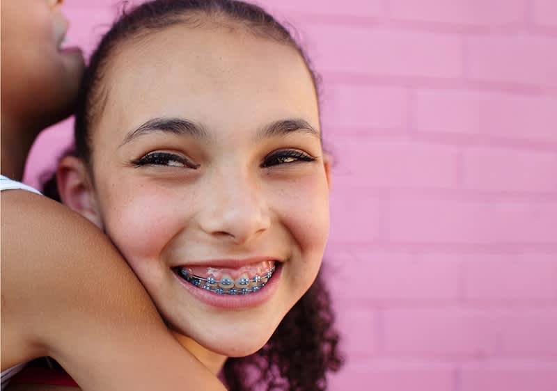 Bambina con apparecchio sorride davanti a un muro rosa mentre viene abbracciata da un'altra persona.