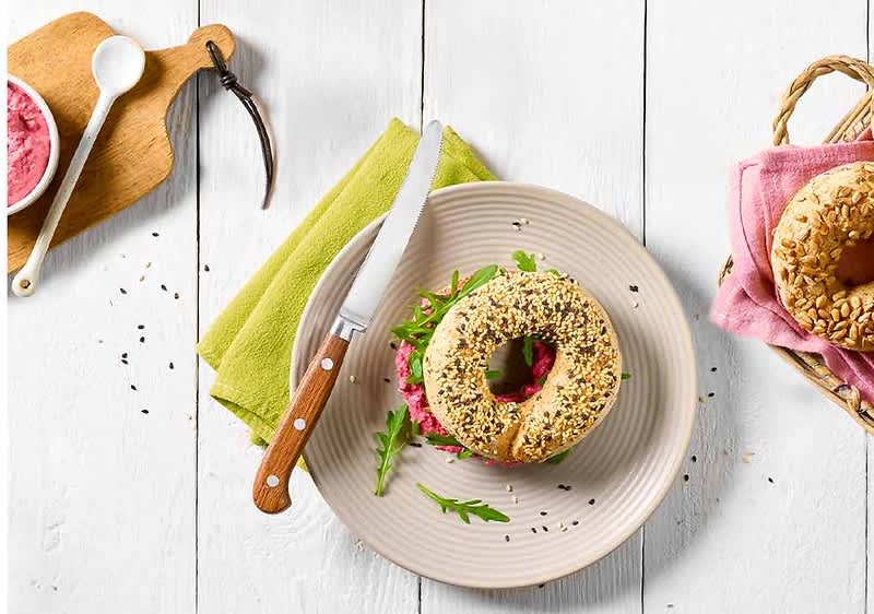 Bagel senza glutine con semi e crema di barbabietola su piatto bianco e coltello