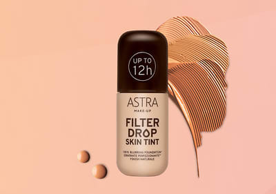 Fondotinta Astra Filter Drop Skin Tint con tappo nero su sfondo arancione con elementi grafici di fondotinta