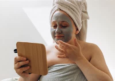 Donna con asciugamano in testa tiene uno specchio in mano e si guarda mentre applica una maschera viso