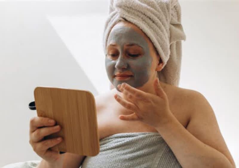 Donna con asciugamano in testa tiene uno specchio in mano e si guarda mentre applica una maschera viso