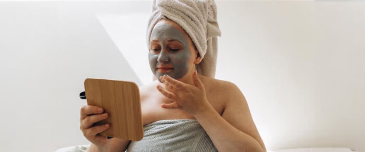 Donna con asciugamano in testa tiene uno specchio in mano e si guarda mentre applica una maschera viso