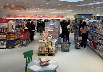 Punto vendita dm affollato con persone che fanno acquisti tra scaffali pieni di prodotti nel reparto Neonati e bambini