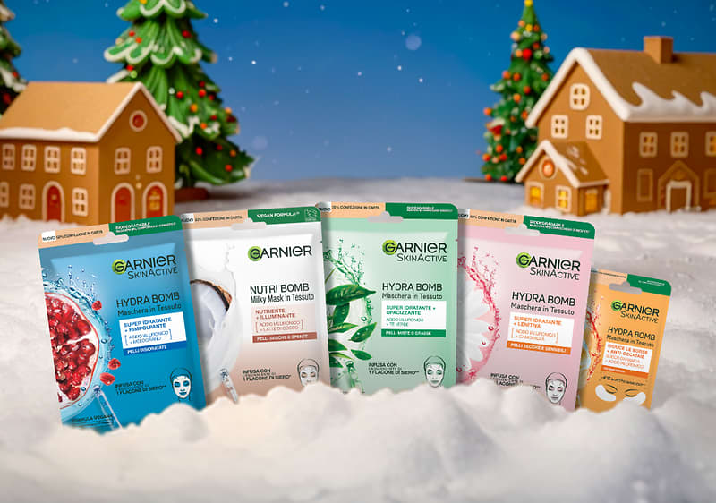 Maschere viso Garnier su neve con case natalizie sullo sfondo