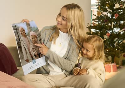 Mamma e figlia guardano insieme calendario con foto di bambini, sullo sfondo albero di Natale decorato.
