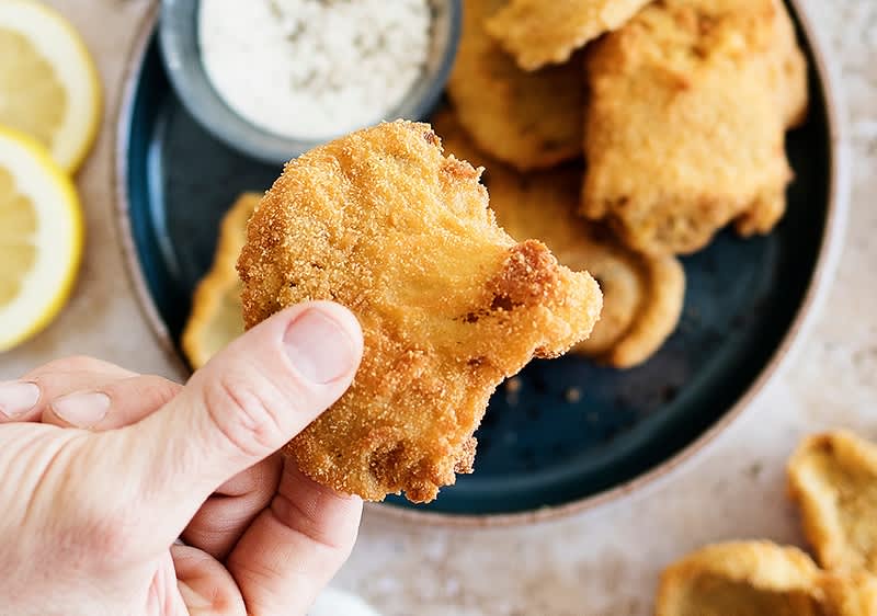 Mano tiene un pezzo di cibo fritto impanato su un piatto con altri pezzi e una salsa.