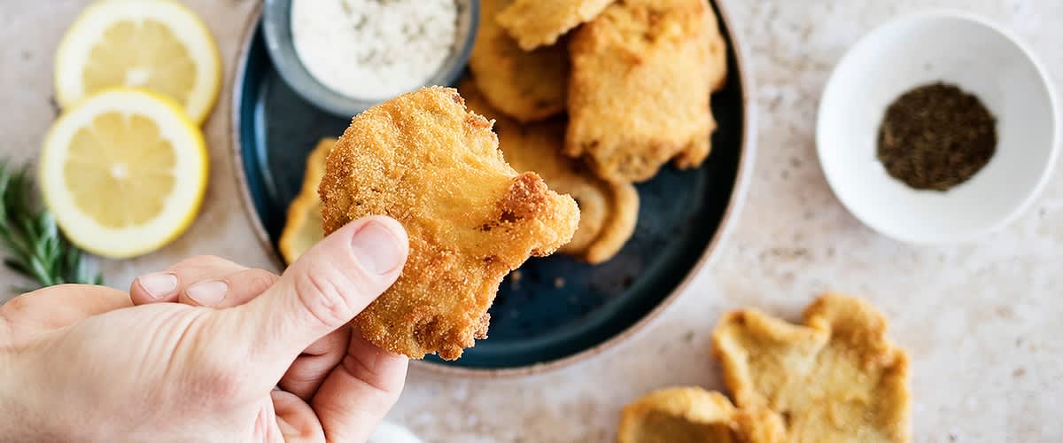 Mano tiene un pezzo di cibo fritto impanato su un piatto con altri pezzi e una salsa.