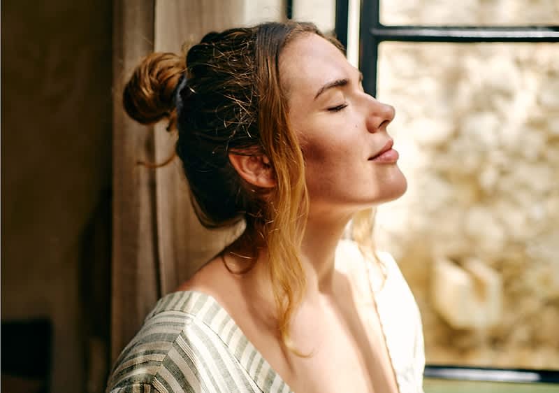 Una donna si trova di fronte ad una finestra e prende il sole con gli occhi chiusi.