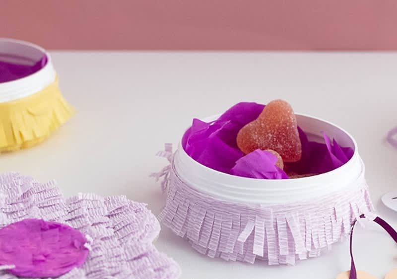 Piccolo vasetto bianco con nastro di frange rosa, riempita con fiorellini di carta viola e una caramella a forma di cuore ricoperta di zucchero.