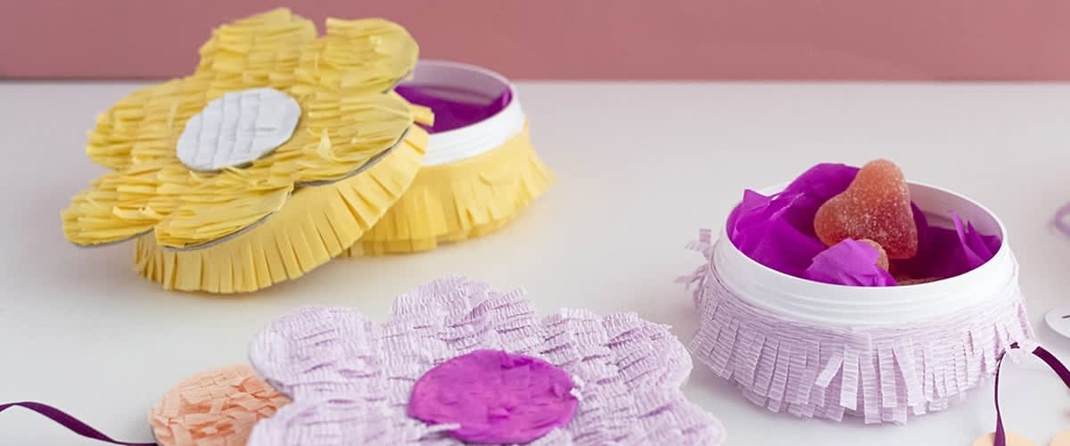Piccolo vasetto bianco con nastro di frange rosa, riempita con fiorellini di carta viola e una caramella a forma di cuore ricoperta di zucchero.