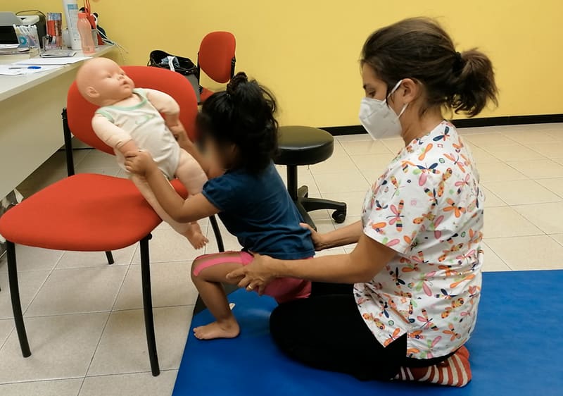 Una bambina accovacciata che tiene un bambolotto appoggiato su una sedia rossa, affiancata da una donna seduta a terra su un tappetino blu che la assiste appoggiandole una mano sulla schiena; la donna indossa una mascherina e una casacca con stampa colorata senza testo evidente