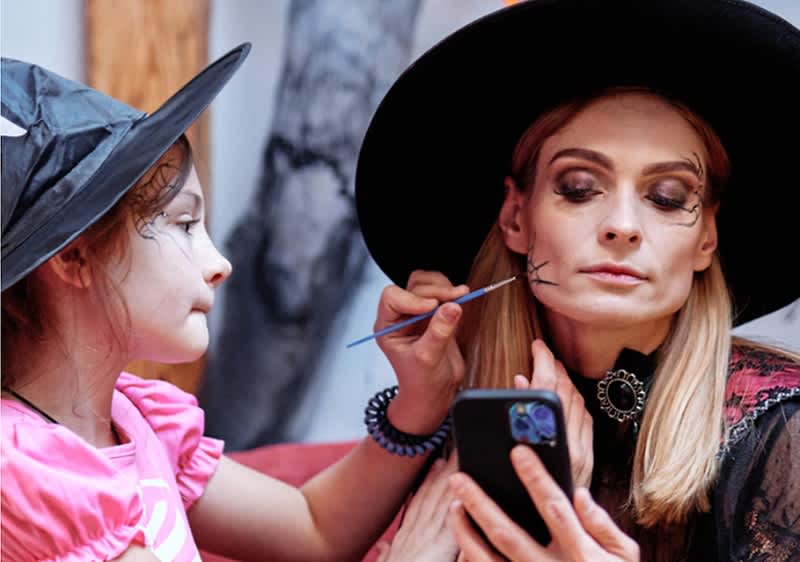 Bambina con cappello da strega che dipinge con un pennello un disegno a ragnatela sul volto di una donna adulta vestita da strega, la donna tiene uno smartphone per specchiarsi mentre la bambina concentra il lavoro sul mento e guancia