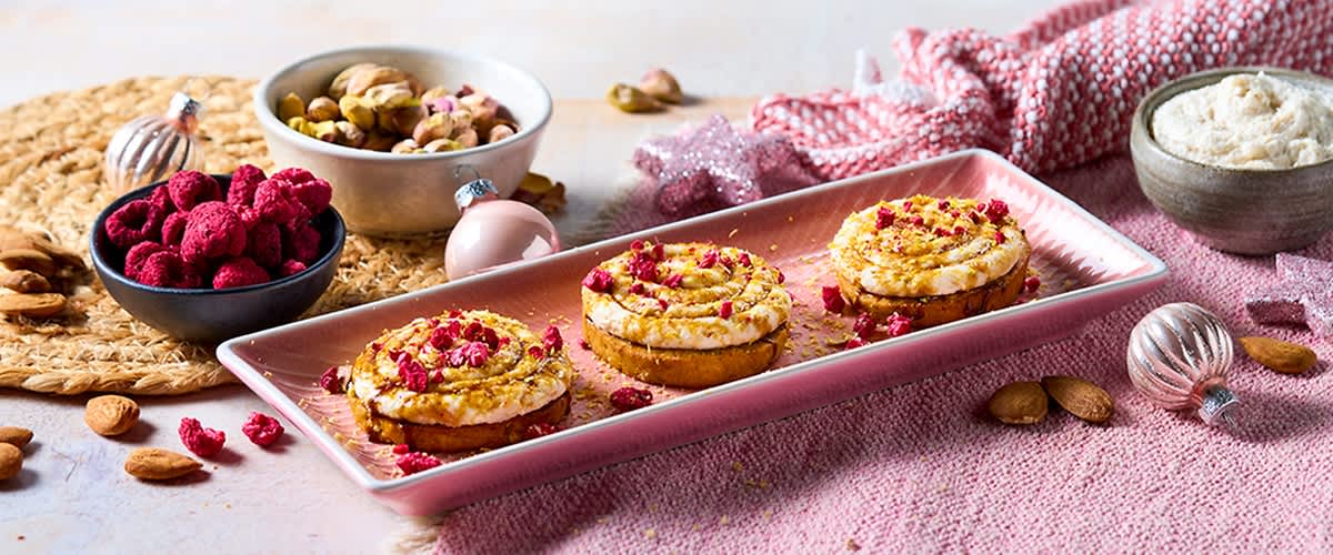 Tre tartine rotonde su un vassoio rosa, ciascuna con base croccante, crema spalmata, granella dorata e pezzi di frutta rossa secca, accanto ciotole con pistacchi e decorazioni natalizie su tovaglia rosa