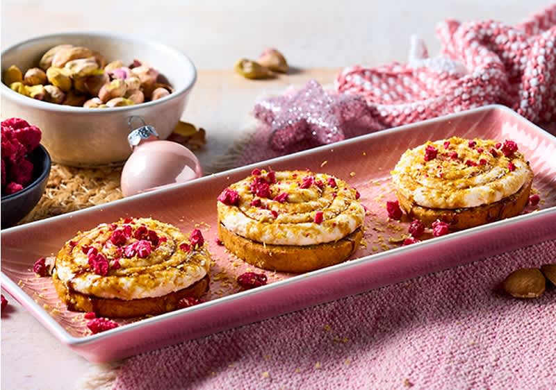 Tre tartine rotonde su un vassoio rosa, ognuna con base croccante, crema bianca spalmata, granella dorata e pezzetti di frutti rossi disidratati; sullo sfondo ciotole con pistacchi e frutti rossi e una pallina decorativa rosa