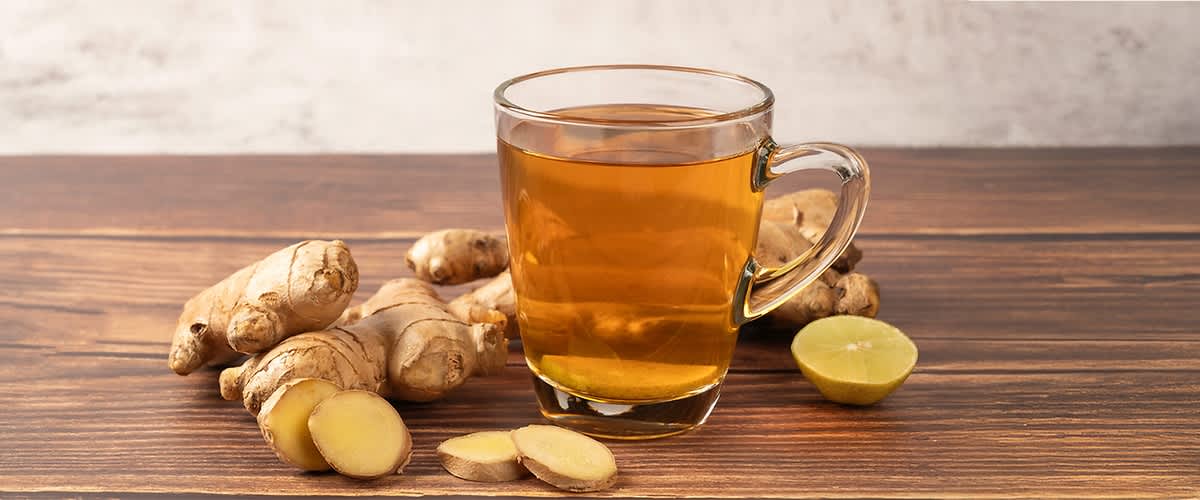Tazza di vetro trasparente con tè allo zenzero su tavolo di legno, circondata da radici di zenzero intere e a fette e metà di lime verde sul lato destro