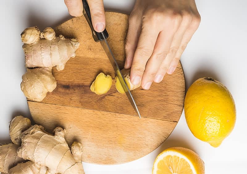 Mani che affettano una radice di zenzero su un tagliere rotondo di legno, con altri pezzi di zenzero a sinistra e due limoni, uno intero e uno tagliato a metà, sul piano bianco