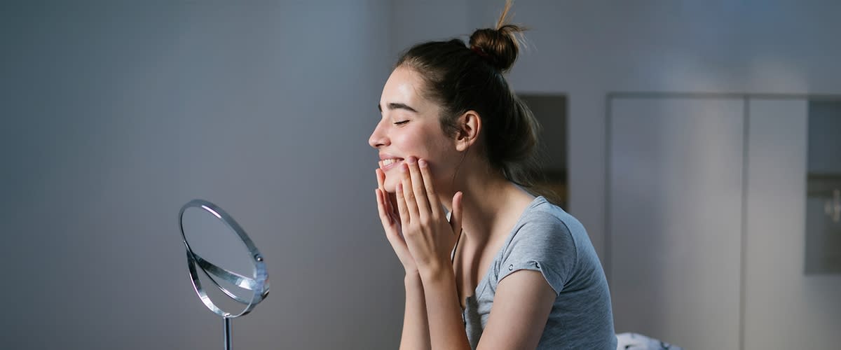 Giovane donna con capelli raccolti a nodo che sorride mentre si tocca il viso davanti a uno specchio tondo, indossa una maglietta grigia a maniche corte e si trova in un interno neutro
