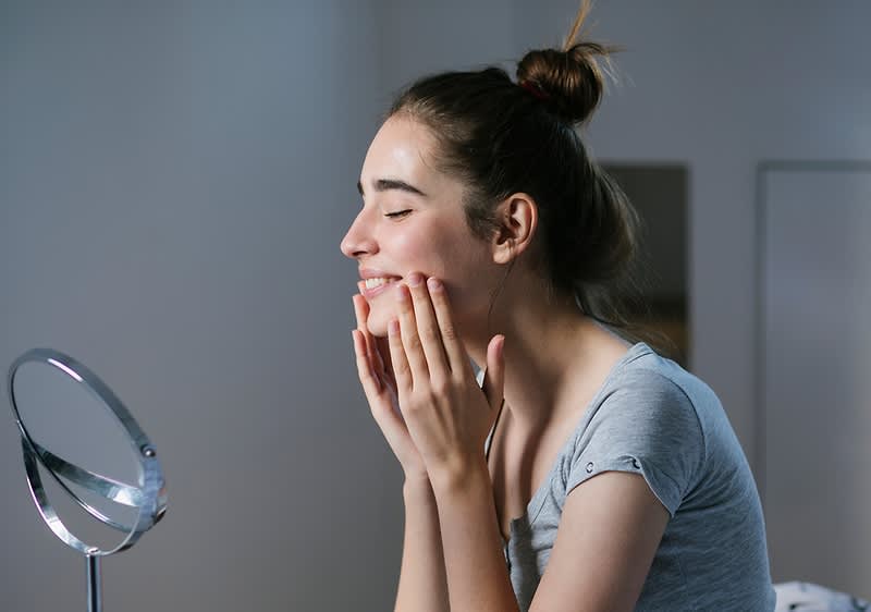 Una giovane donna con capelli raccolti a chignon sorride davanti a uno specchio mentre si tocca delicatamente il viso con entrambe le mani, indossando una maglietta grigia a maniche corte