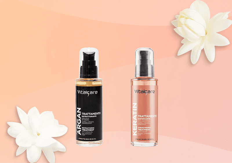 Due flaconi con erogatore del marchio Vitalcare su sfondo sfumato pescato: a sinistra olio per capelli Argan in bottiglia nera con etichetta verticale bianca con scritto ARGAN e dicitura TRATTAMENTO, a destra flacone rosa/ambra etichettato KERATIN con dicitura TRATTAMENTO, decorati con due fiori bianchi