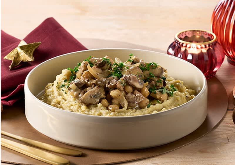 Piatto rotondo con polenta cremosa sormontata da funghi e ceci saltati, guarniti con prezzemolo tritato; a sinistra tovagliolo bordeaux con stella dorata, posate dorate in primo piano e candele decorative rosse sullo sfondo