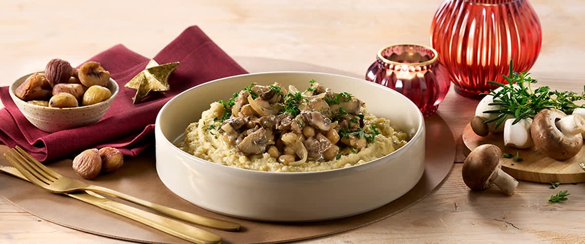 Piatto rotondo bianco con purea di patate cremosa sormontata da funghi saltati e prezzemolo tritato, posato su sottopiatto dorato; a sinistra tovagliolo bordeaux con stella dorata e posate dorate, a destra due portacandele decorativi rossi