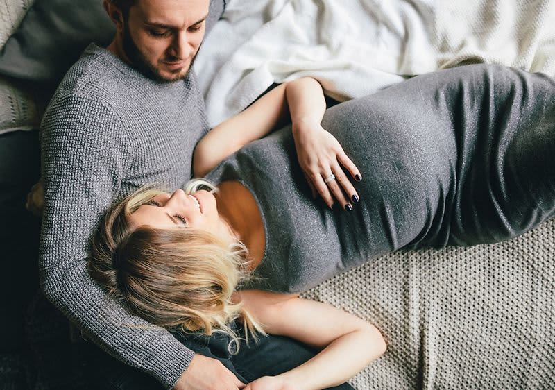 Mężczyzna leży za kobietą w ciąży, oboje trzymają dłonie na brzuchu ciążowym