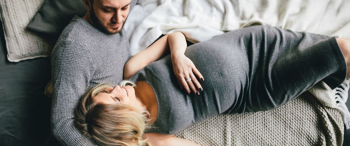 Mężczyzna leży za kobietą w ciąży, oboje trzymają dłonie na brzuchu ciążowym