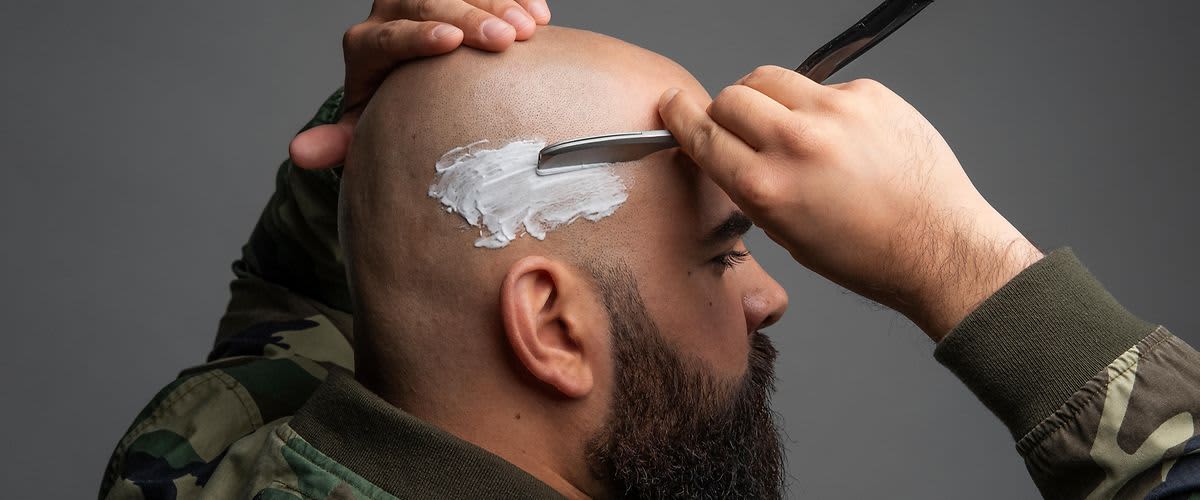 Mężczyzna z długą brodą przytrzymuje brzytwę przy wygolonej na łyso głowie.