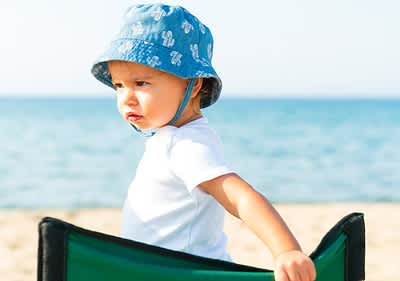 Dziecko w niebieskim kapeluszu siedzi na plaży na zielonym krześle, w tle morze i niebo