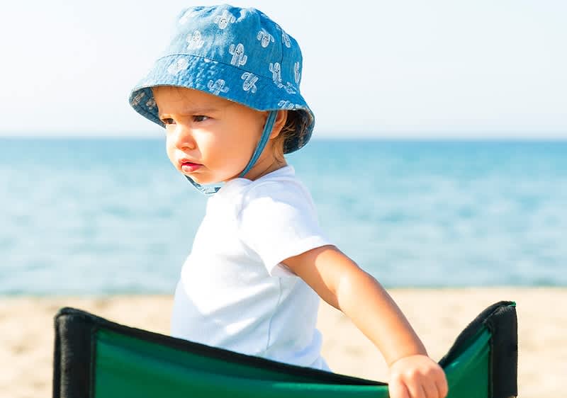 Dziecko w niebieskim kapeluszu siedzi na plaży na zielonym krześle, w tle morze i niebo