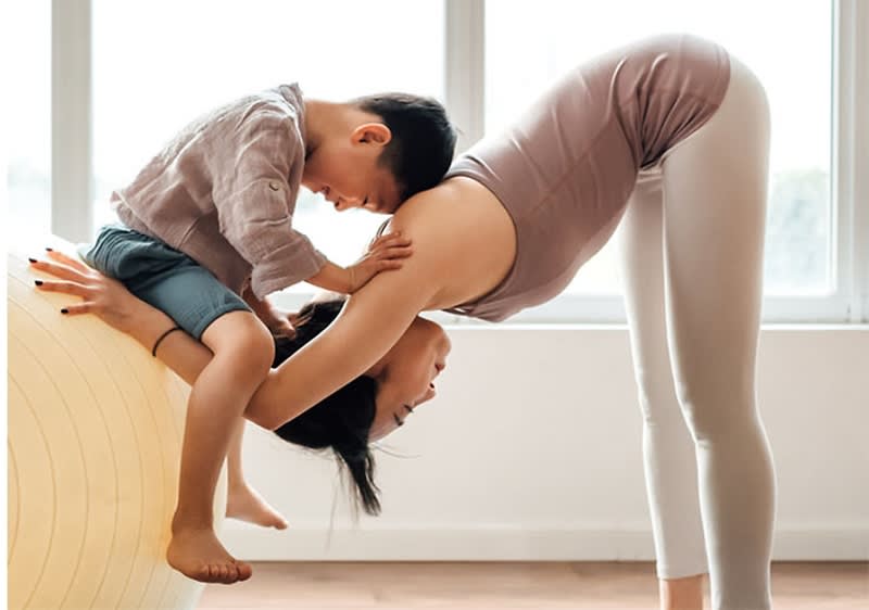 Kobieta i chłopiec ćwiczą razem, kobieta pochyla się do przodu, chłopiec dotyka jej pleców, w jasnym pomieszczeniu z dużym piłką do ćwiczeń