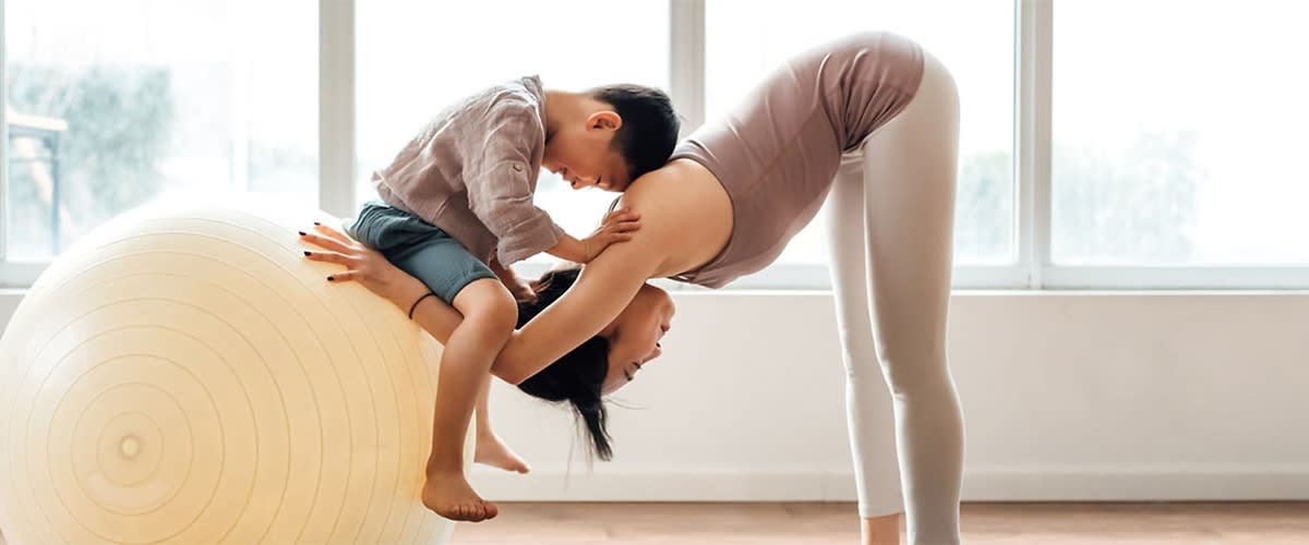 Kobieta i chłopiec ćwiczą razem, kobieta pochyla się do przodu, chłopiec dotyka jej pleców, w jasnym pomieszczeniu z dużym piłką do ćwiczeń