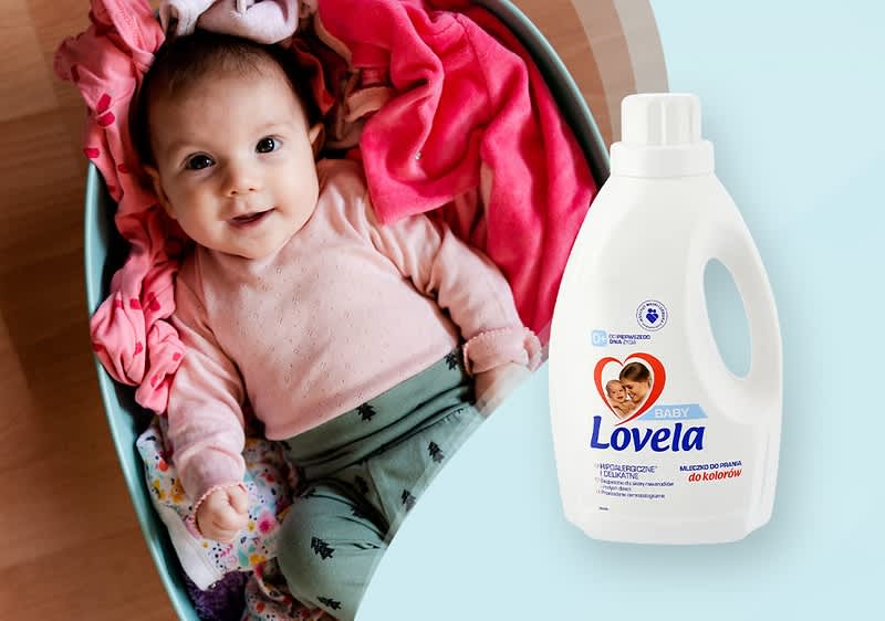Butelka płynu do prania Lovela na drewnianej powierzchni obok kolorowych ubranek w koszu, w którm znajduje się niemowlę.