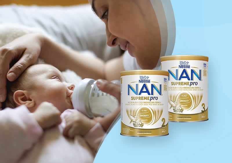 Niemowlę leżące na białym prześcieradle, trzymające butelkę, obok dwie puszki mleka NAN
