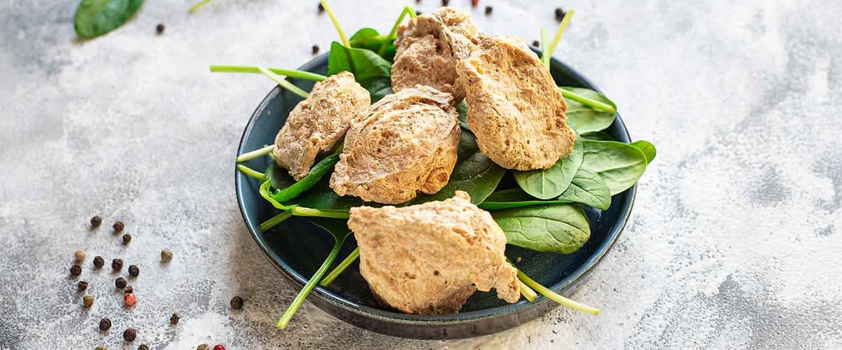 Na ciemnym talerzu leży kilka kawałków brązowego, przypominającego mięso seitanu na świeżych zielonych liściach szpinaku, otoczonych rozrzuconymi czarnymi i czerwonymi ziarnami pieprzu na jasnej kamiennej powierzchni.