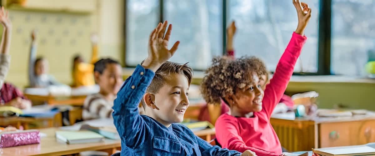 Dwoje dzieci w klasie podnosi ręce, żeby się zgłosić, siedząc przy drewnianych ławkach