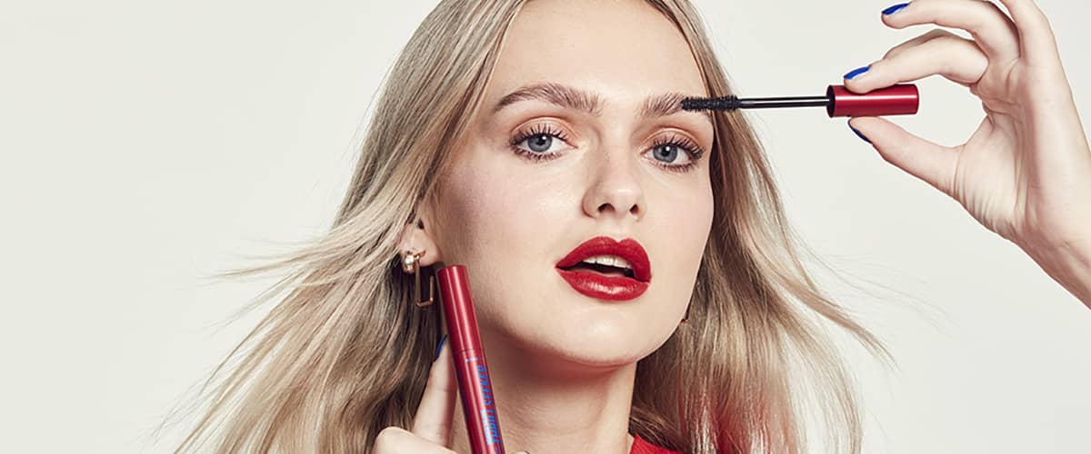 Femeie aplicând mascara pe gene cu păr blond și cercei aurii