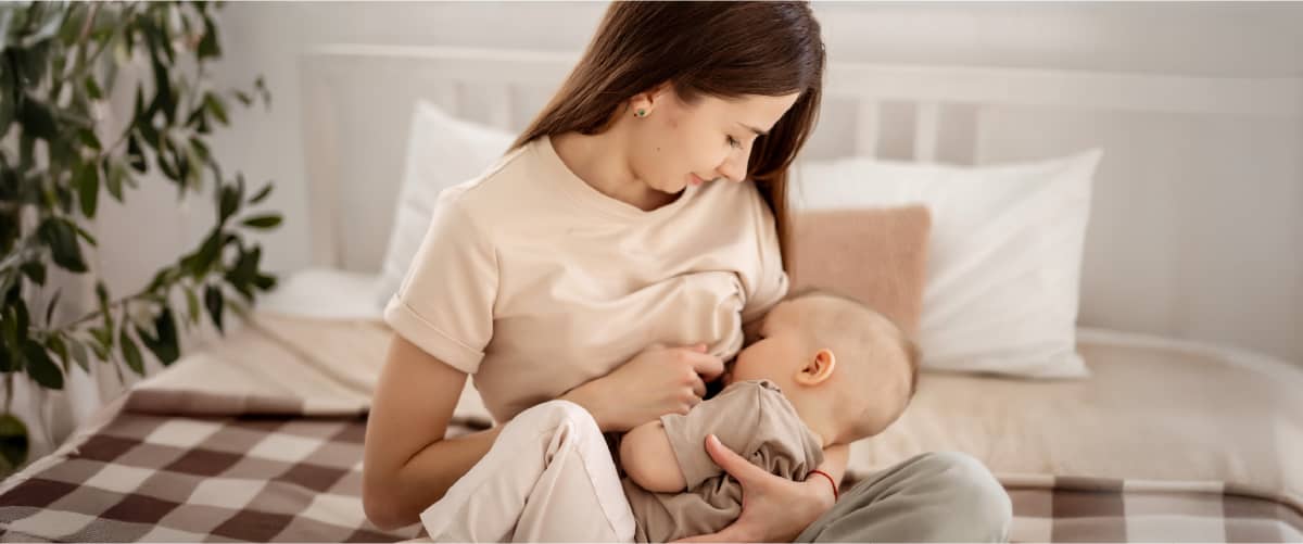 Mamă alăptând un bebeluș pe pat într-o cameră luminoasă