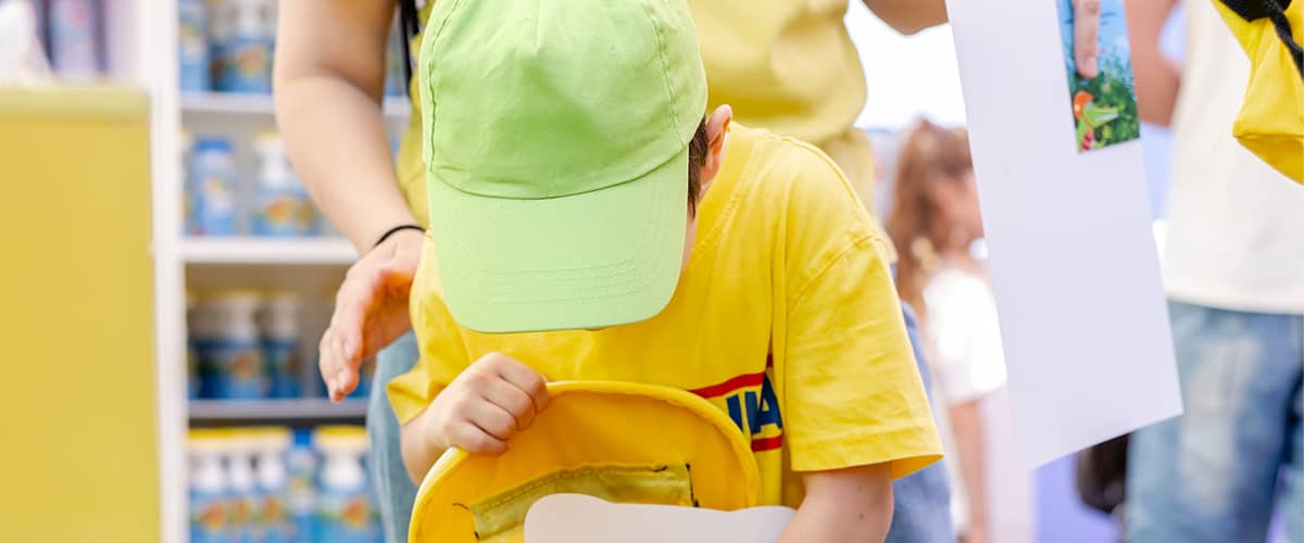 copil în tricou galben și șapcă verde căutând în ghiozdan într-un magazin