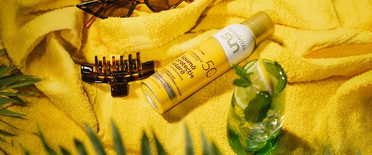 cremă cu protecție solară SPF50 pe prosop galben lângă ochelari, clemă de păr și pahar cu băutură și frunze