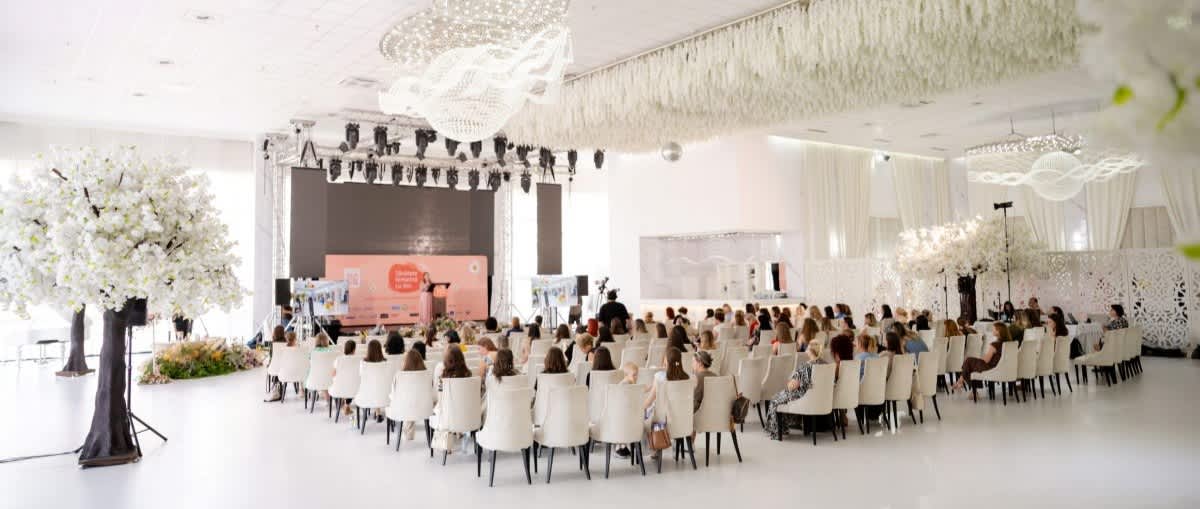 Eveniment într-o sală luminoasă cu public așezat pe scaune, scenă și decoruri florale