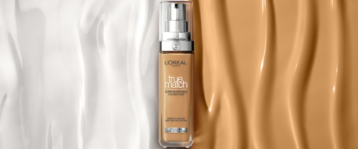 Sticlă transparentă de fond de ten L'Oréal True Match cu pompă argintie, plasată vertical pe un fundal cu textura fluidă albă în stânga și nuanță maro-bej în dreapta, textul de pe ambalaj „true match” vizibil