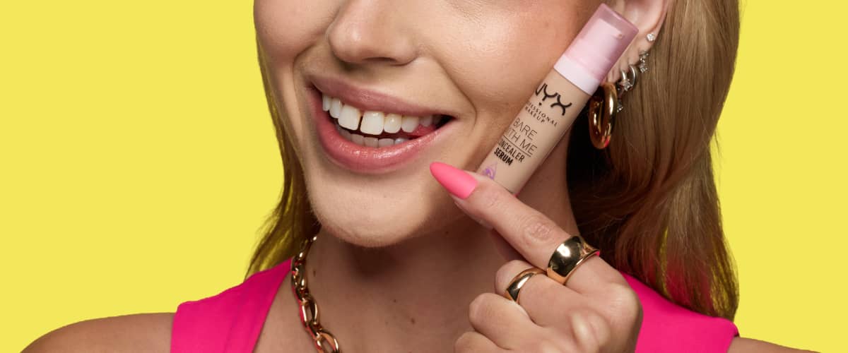 O femeie zâmbitoare ține aproape de obraz un concealer lichid într-un recipient bej cu capac roz, poartă o bluză roz, inele aurii și lanț gros aurit la gât, unghiile lungi roz neon și cercei multiple în urechea dreaptă