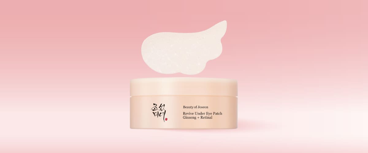 Recipientor rotund crem cu capac, pe fundal roz pal, etichetă cu text „Beauty of Joseon Revive Under Eye Patch Ginseng + Retinal” și un patch alb translucid în formă de semilună plasat deasupra recipientului