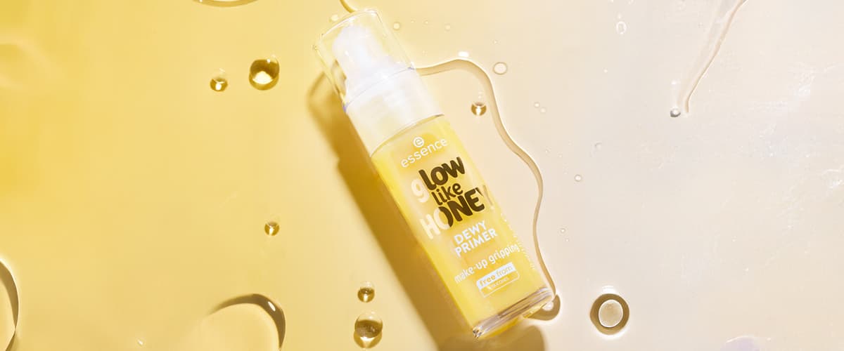 Sticlă transparentă cu pompă de primer pentru față „glow like HONEY” marca essence, lichid galben în interior, etichetă galbenă cu text alb și negru „Dewy Primer” pe fundal deschis cu picături de lichid și reflexii luminoase